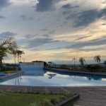 Una serena piscina al aire libre refleja un cielo nublado al amanecer o al atardecer, rodeada de palmeras y zonas verdes ajardinadas. Al fondo, un cartel indica Pereira, cerca de la famosa Biblioteca Pública Hugo Ángel Jaramillo.
