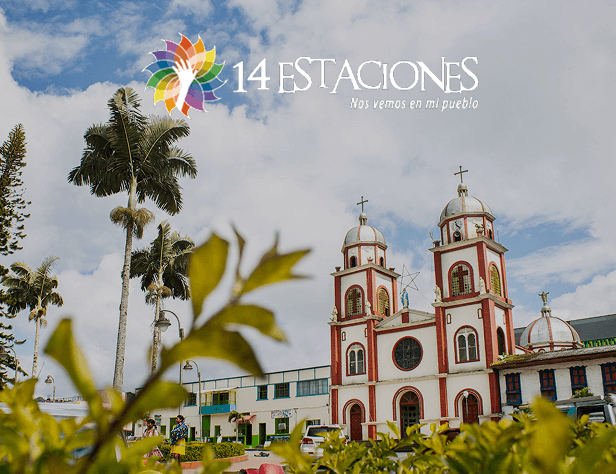 Una colorida iglesia con campanarios gemelos se alza detrás de plantas verdes en Pueblo Rico, Risaralda. El cielo está nublado. En la parte superior, un logotipo multicolor y un texto rezan: 14 ESTACIONES Nos vemos en mi pueblo.