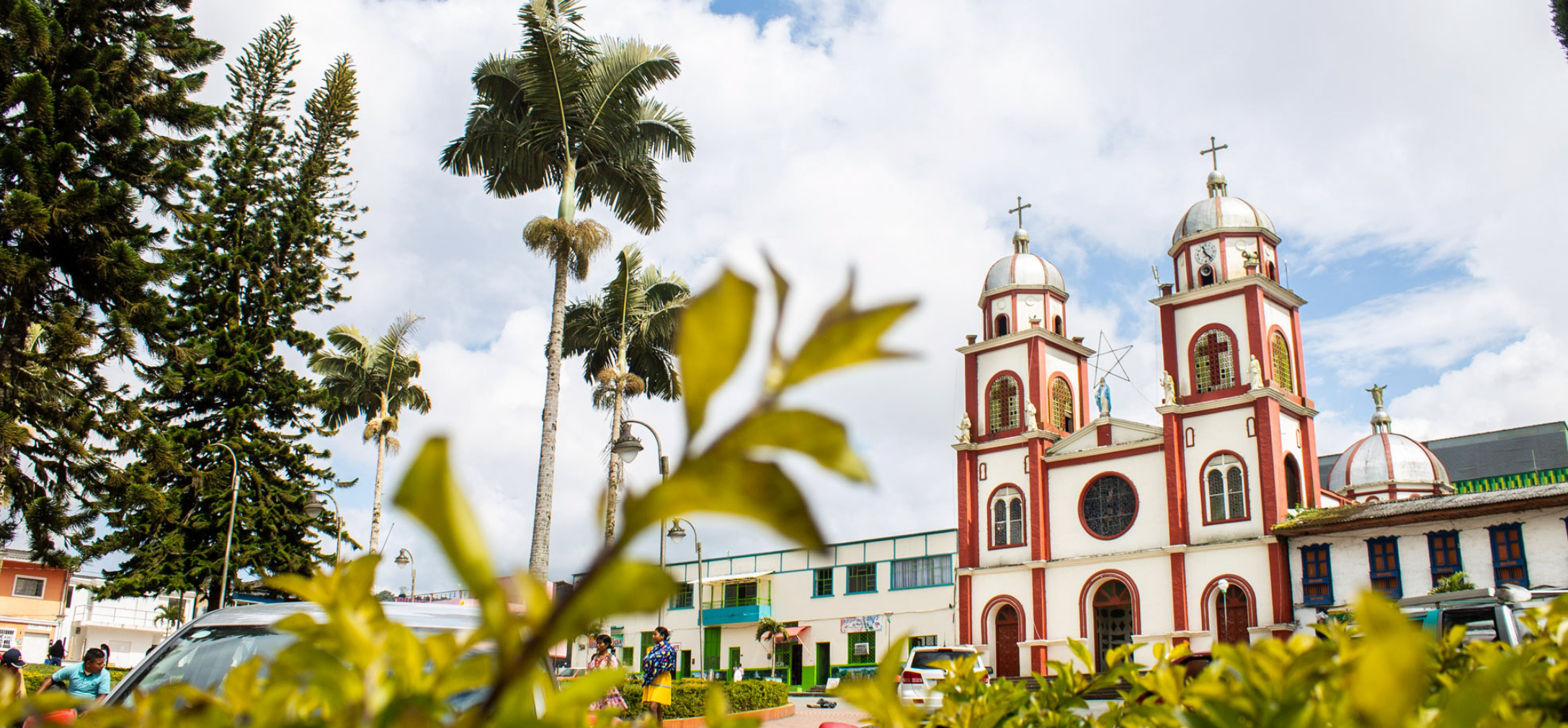 Altas palmeras y vegetación enmarcan una iglesia roja y blanca con dos torres bajo un cielo parcialmente nublado, a pocos pasos de la vibrante Biblioteca Municipal en la animada plaza de la ciudad.