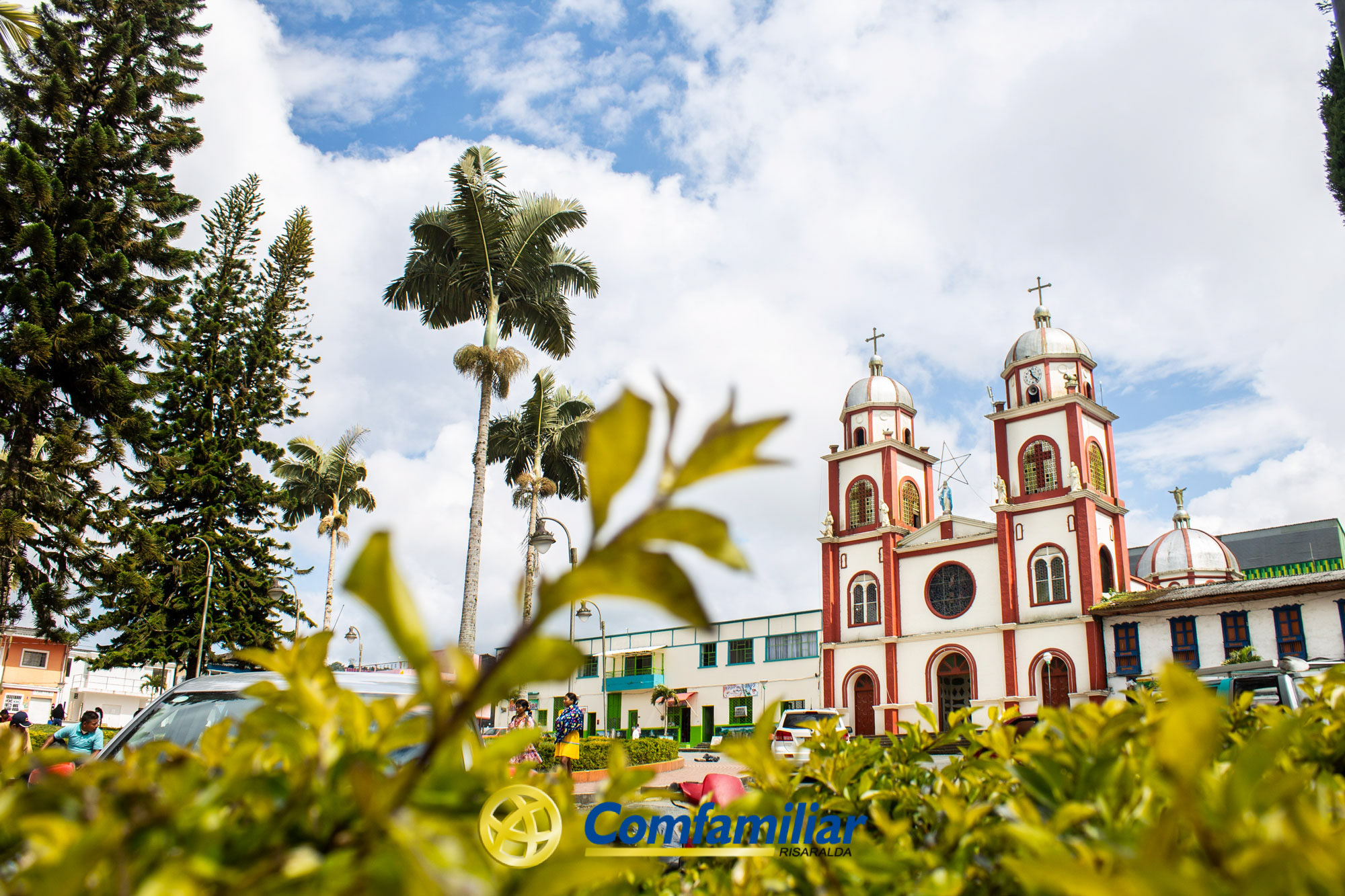 Una foto vibrante de la iglesia roja y blanca con dos torres en el Corregimiento de Santa Cecilia, rodeada de palmeras y vegetación bajo un cielo parcialmente nublado. En primer plano, plantas frondosas, la Biblioteca Pública y el logotipo amarillo de Comfamiliar.