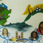 Un colorido mural con un mapa verde con Pueblo Rico Risaralda, una bandera colombiana, tres mujeres de diversos orígenes, la iglesia de Santa Cecilia, montañas y remolinos decorativos azules sobre una pared blanca.
