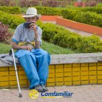 Un anciano con sombrero blanco y pantalones azules se sienta en un banco del parque, cerca de unos vibrantes parterres, con un teléfono en la mano. Un bastón descansa a su lado. El logotipo "Comfamiliar Risaralda" aparece en la parte inferior, destacando la Biblioteca Pública en el Corregimiento de Santa Cecilia.