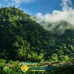 Una exuberante montaña verde cubierta de un denso bosque con nubes bajas se eleva sobre Santa Cecilia. En la base se ven pequeñas casas de colores y una Biblioteca Pública. El logotipo de Confamiliar aparece en la parte inferior central de la imagen.