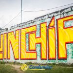 Mural grande y colorido en una pared con la palabra Quinchía en letras amarillas y rojas. Debajo, aparece el logotipo de Comfamiliar Risaralda y una mención a la Biblioteca Municipal. Al fondo se ven árboles verdes y montañas.