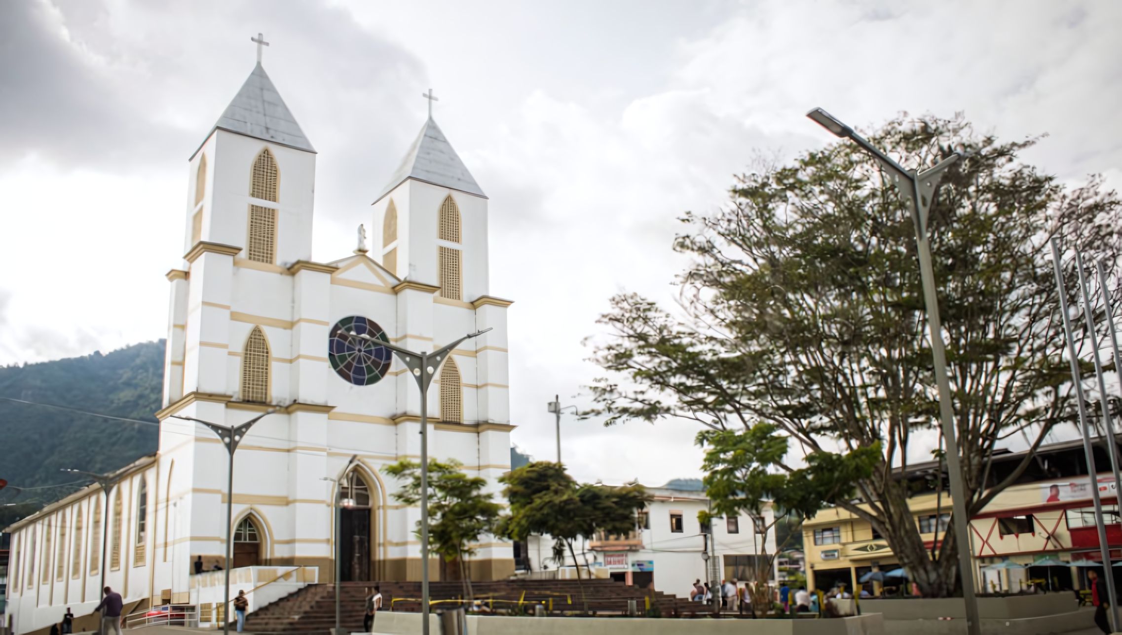 Una iglesia blanca con campanarios gemelos y una gran vidriera circular se alza en una plaza municipal, cerca de la Biblioteca Pública, rodeada de árboles, edificios y gente bajo un cielo nublado.