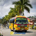 Un autobús de colores con la etiqueta Batero circula por una calle bordeada de palmeras en una pequeña ciudad bajo un cielo nublado, pasando junto a casas y edificios. Abajo aparece el logotipo de Comfamiliar, junto a pancartas de la Biblioteca Pública Danilo Calamata.