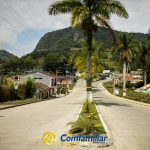 Una calle soleada bordeada de palmeras conduce a una pequeña ciudad rodeada de verdes montañas. A la derecha se ven casas, murales y la Biblioteca Municipal, con el logotipo "Comfamiliar" en la parte inferior de la imagen.