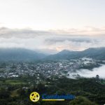 Vista panorámica de un municipio rodeado de verdes montañas, con nubes bajas y niebla que atraviesan el paisaje. El cielo está nublado y el logotipo de Comfamiliar aparece en la parte inferior central, cerca de la Biblioteca Pública.