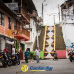 Una escena callejera en un barrio montañoso con gente, motocicletas, edificios de colores y la vibrante escalera de mosaico cerca de la Biblioteca Pública Danilo Calamata. El logotipo de Comfamiliar aparece en la parte inferior central de la imagen.