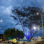 Un parque público al atardecer con farolas modernas, un árbol alto iluminado con luces azules, cielo nublado y el logotipo de Comfamiliar en la parte inferior central. Cerca de bancos y vegetación, la gente disfruta del ambiente junto a la Biblioteca Pública Municipal.