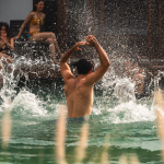 Un hombre chapotea juguetonamente en una piscina de Termales San Vicente, de espaldas a la cámara. Al fondo, dos mujeres sentadas al borde de la piscina, sonríen y observan la acción mientras la luz del sol resalta la enérgica escena.