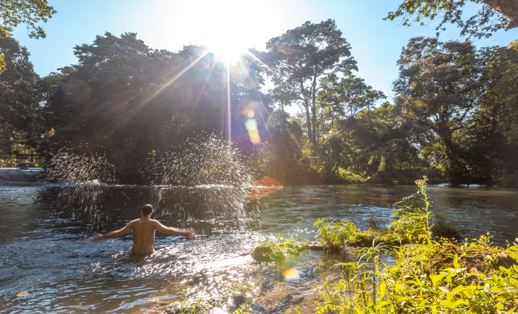Una persona se sumerge hasta la cintura en un río de los Termales de San Vicente, chapoteando bajo la luz del sol. Los árboles y la densa vegetación rodean el río, y la luz del sol se cuela entre las hojas, creando una escena tranquila y natural.