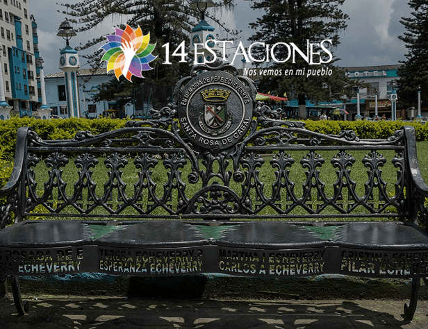 Un banco de metal ornamentado con nombres grabados se encuentra en una plaza de Santa Rosa de Cabal, con edificios, árboles y una torre del reloj detrás. Un colorido logotipo y el texto 14 Estaciones. Nos vemos en mi pueblo aparecen en la parte superior de la imagen.