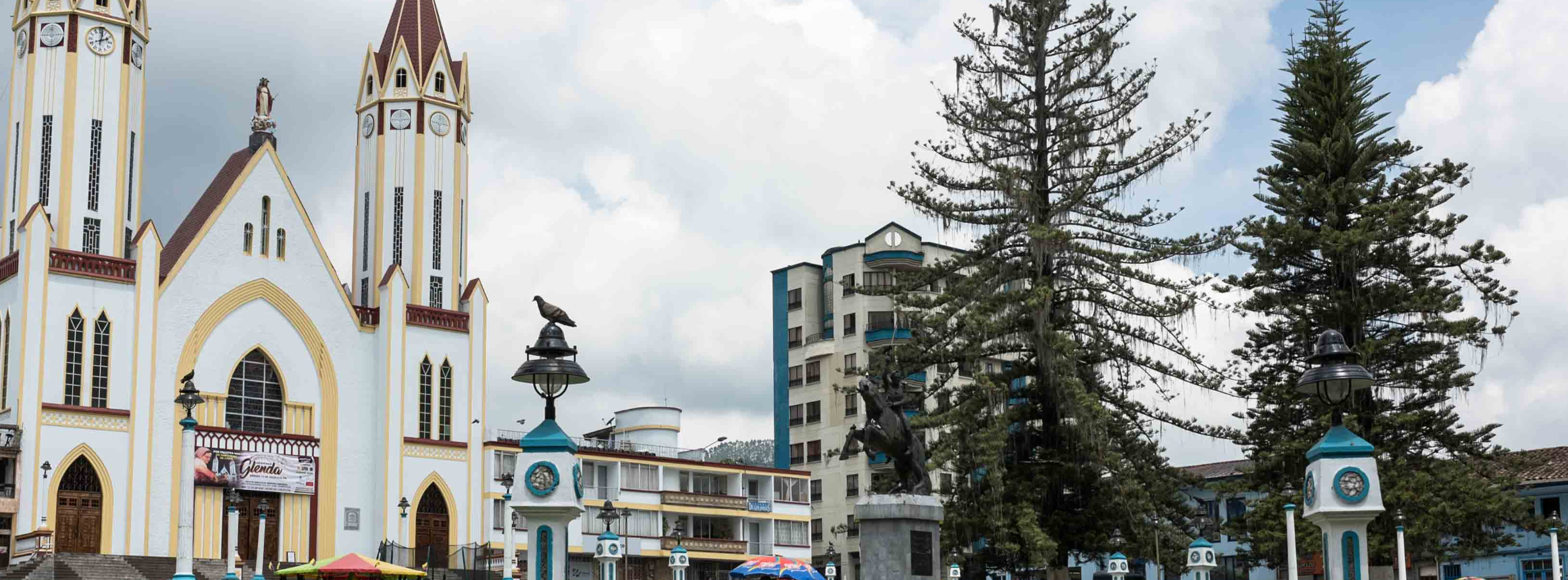 Una gran iglesia con altas torres gemelas se alza junto a una plaza con árboles, farolas, una estatua y la cercana Biblioteca Pública Jaime Fernández Botero bajo un cielo nublado.
