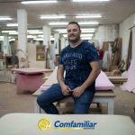 Un hombre con camiseta azul está sentado en un banco de trabajo en un taller de carpintería, sonriendo a la cámara. En primer plano se ve el logotipo de Comfamiliar Risaralda. Herramientas y madera le rodean, evocando el espíritu de las iniciativas de la Biblioteca Pública de Jaime Fernández Botero.