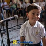 Un niño con un polo blanco sonríe delante de un micrófono en la Biblioteca Pública Jaime Fernández Botero, con otros niños con camisetas similares sentados detrás de él. El logotipo de Comfamiliar aparece en la parte inferior de la imagen.