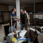 Dos hombres se dan la mano mientras una mujer sentada trabaja con una máquina de coser en un taller lleno de telas y muebles. El logotipo de Comfamiliar aparece en la parte inferior, destacando el soporte inspirado en Jaime Fernández Botero.