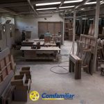 Un taller de carpintería con varios trabajadores montando piezas de mobiliario de madera para la Biblioteca Pública. Varias sillas y piezas de madera están organizadas alrededor de la sala. El logo de Comfamiliar Risaralda es visible en la parte inferior central de la imagen.