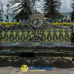 Banco ornamentado de metal negro con nombres grabados y un escudo en el respaldo, situado en un parque cerca de arbustos verdes, edificios y la torre del reloj azul y blanco de la Biblioteca Pública Jaime Fernández Botero. El logotipo de Confamiliar aparece en el pavimento.