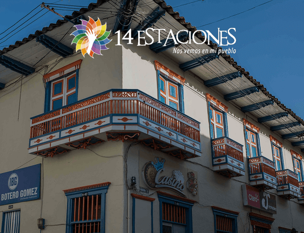 Un tradicional edificio Santuario con coloridos balcones azules y naranjas se asienta bajo un letrero que dice "14 Estaciones, Nos vemos en mi pueblo." Varios letreros de negocios se ven en la planta baja de Risaralda.