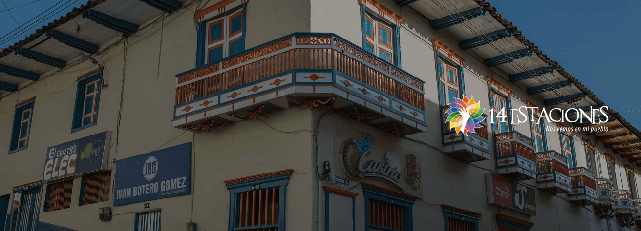 Edificio de estilo colonial con coloridos balcones en la pequeña ciudad de Santuario, Risaralda, con letreros de tiendas a nivel de la calle y el logotipo de 14 Estaciones con un vibrante diseño de flores en la esquina superior derecha.