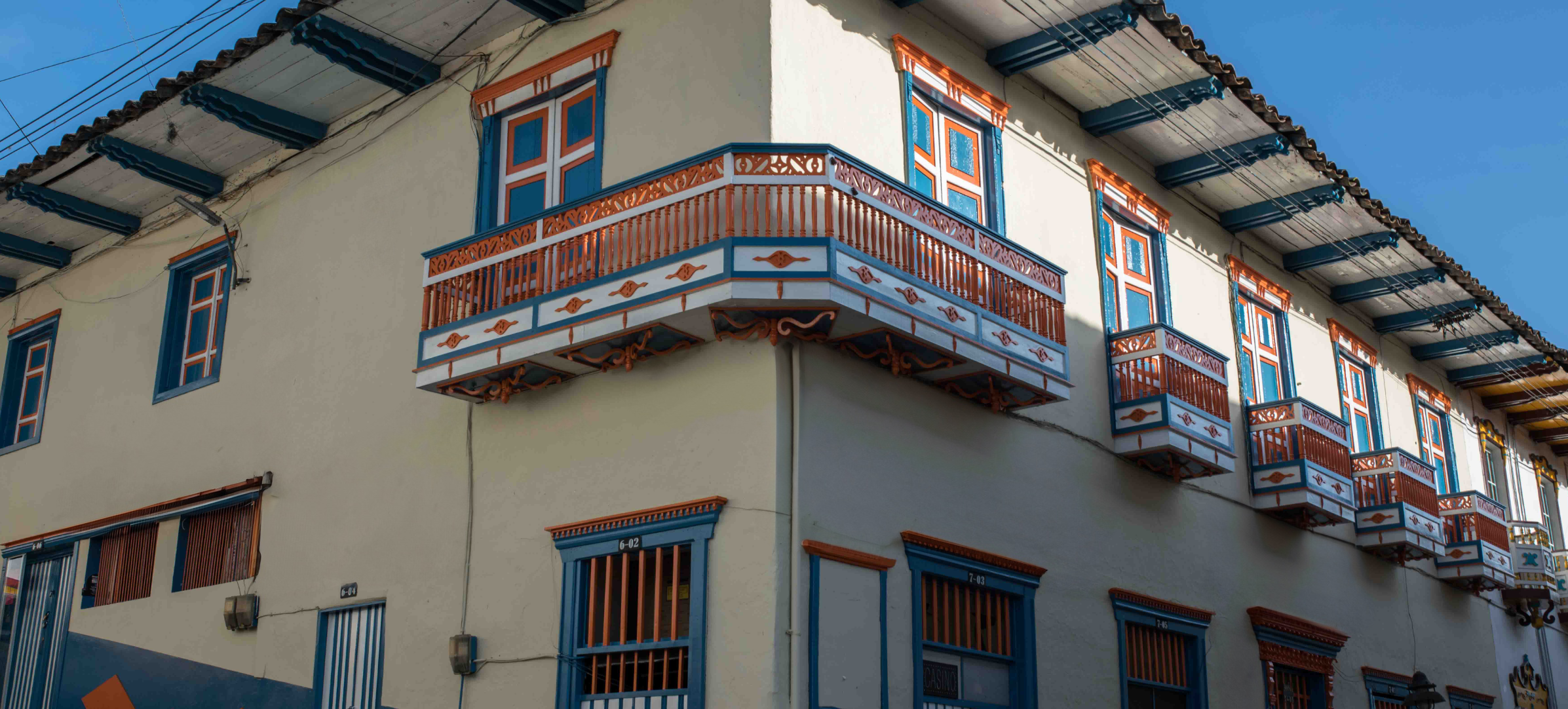 Un edificio tradicional de estilo colonial con balcones de madera ornamentados, adornos azules y naranjas y barandillas decorativas se alza orgulloso en Santuario contra un cielo azul despejado.