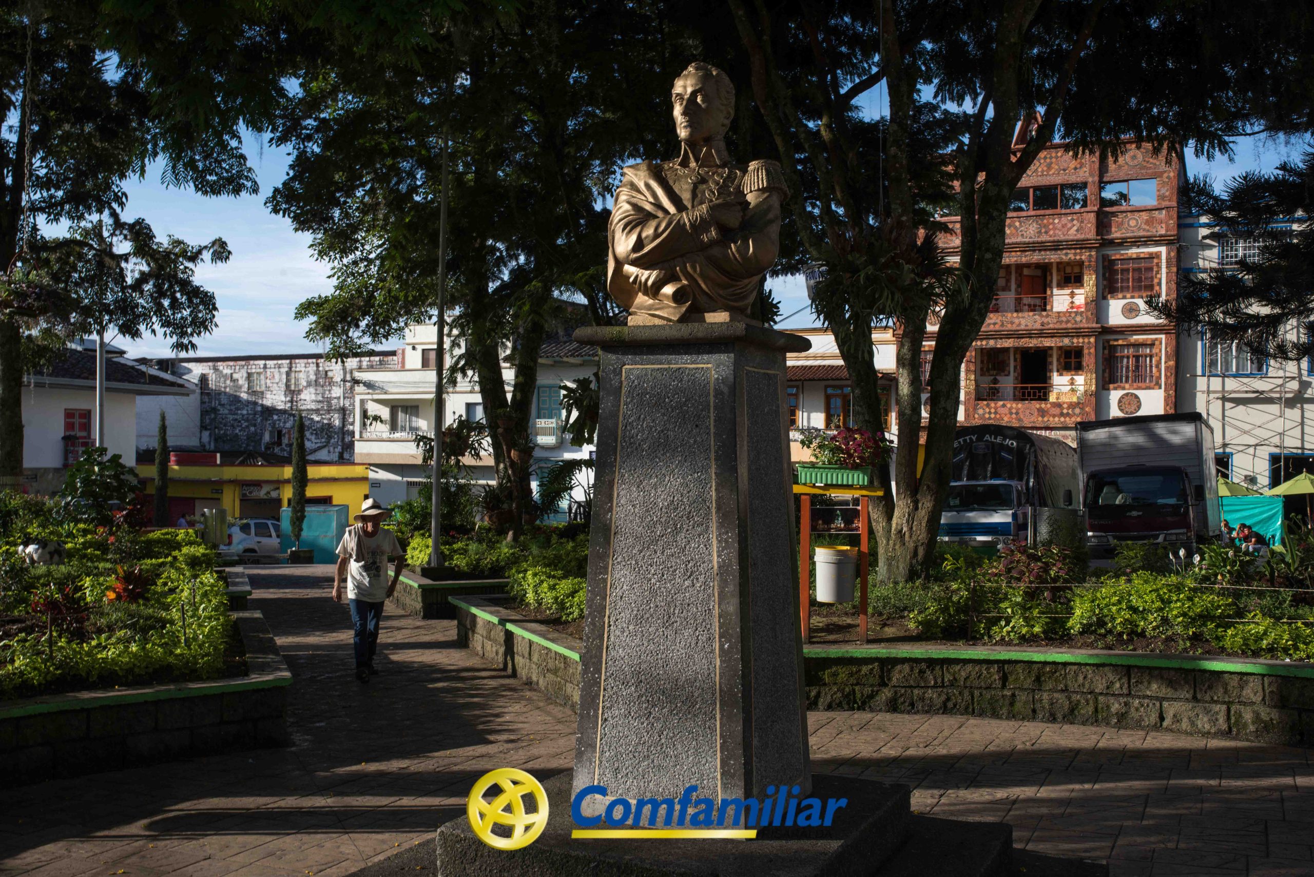 Un busto de bronce de una persona sobre un pedestal de piedra se encuentra en una plaza sombreada cerca de la Biblioteca Pública Comfamiliar Santuario, con árboles, gente caminando y edificios en el fondo. La palabra Comfamiliar es visible en la parte inferior.