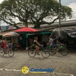 Cuatro personas pasan en bicicleta por delante de puestos de mercado cubiertos con lonas y paraguas en un día nublado. Al fondo, un gran árbol y la Biblioteca Pública Municipal Francisco Jaramillo Ochoa, con el logotipo de Comfamiliar en la parte inferior de la imagen.