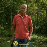 Un hombre mayor vestido con un polo rojo se encuentra en un frondoso y verde bosque cerca de la Biblioteca Pública. La luz del sol se filtra entre los árboles. El logotipo y la palabra Comfamiliar aparecen en la parte inferior de la imagen.