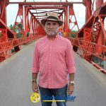 Un hombre mayor con camisa rosa y sombrero está de pie sobre un puente de metal rojo, mirando a la cámara. A la izquierda se ve parcialmente una rueda de bicicleta. En la parte inferior aparece el logotipo de la Biblioteca Pública Municipal Francisco Jaramillo Ochoa.
