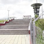 Unos anchos escalones de hormigón conducen a una plataforma con barandilla metálica. Una valla de alambre de espino recorre un camino pavimentado junto a la Biblioteca Pública, donde una persona camina a lo lejos bajo un cielo nublado. Fotografía de Hugo Ángel Jaramillo.
