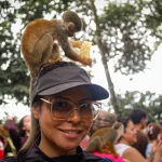 Una mujer con gafas y visera sonríe mientras un pequeño mono se sienta en su cabeza a comer. Otro mono está sobre su hombro. En el fondo se ven personas y árboles.