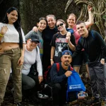 Un grupo de ocho personas sonríen y posan juntas en una zona boscosa cerca de grandes raíces de árboles. Visten de manera informal y parecen estar disfrutando de una aventura al aire libre o de una excursión.