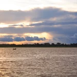 Una pequeña embarcación flota en un ancho río bajo un cielo dramático con la luz del sol asomando entre espesas nubes cerca del horizonte, reflejándose en la superficie del agua.