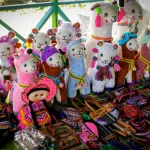 Un colorido puesto de mercado exhibe llamas disecadas hechas a mano, una muñeca con ropa tradicional, bolsos tejidos, cinturones y diversas artesanías textiles, todo ricamente decorado con motivos brillantes y colores vibrantes.