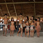 Un grupo de indígenas con atuendos tradicionales y pinturas corporales forman una fila bajo un tejado de paja, algunos con cestas en la mano, y bailan o se preparan para una danza o ceremonia cultural.