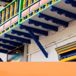 Coloridos edificios de estilo colonial con balcones y elementos decorativos se alinean en una calle bajo un cielo parcialmente nublado. La Biblioteca Pública Isaías Naranjo forma parte de esta vibrante escena. La palabra Apía aparece en un gran texto blanco en la esquina inferior derecha.