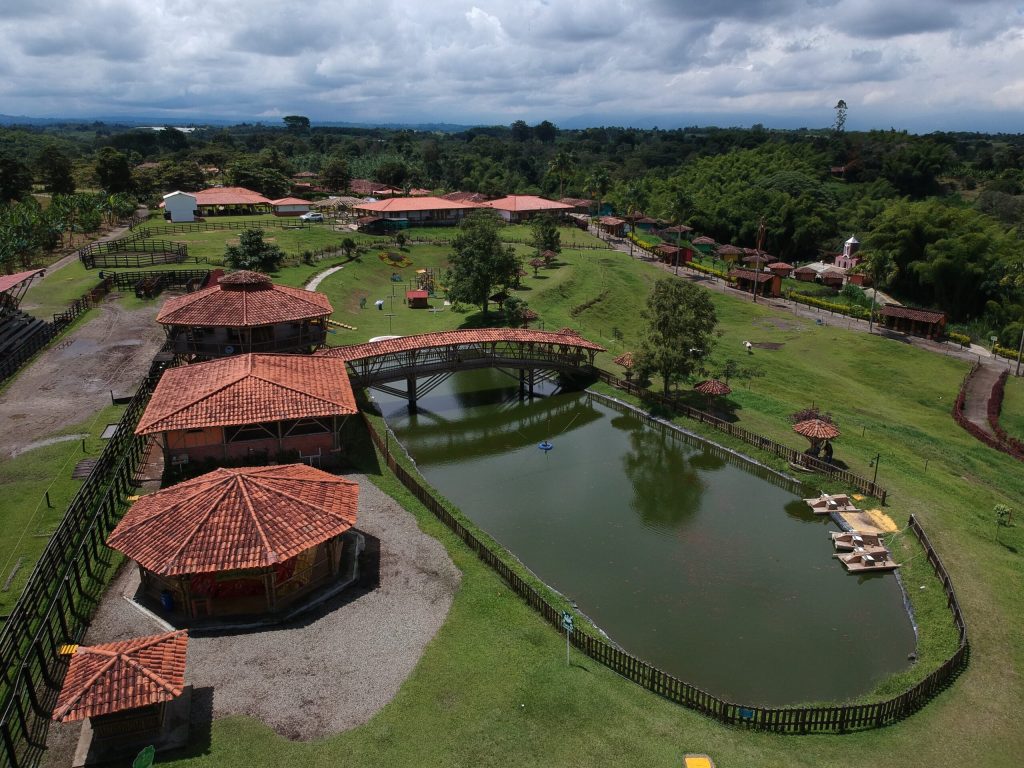 Vista aérea del Parque los arrieros, un parque rural con edificios de tejados rojos, un puente peatonal sobre un estanque, césped verde, árboles y un cielo nublado al fondo. Caminos y vallas dividen el terreno ajardinado.