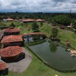 Vista aérea del Parque los arrieros, un parque rural con edificios de tejados rojos, un puente peatonal sobre un estanque, césped verde, árboles y un cielo nublado al fondo. Caminos y vallas dividen el terreno ajardinado.
