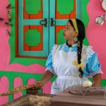 Una mujer con vestido azul y delantal blanco está de pie en una colorida cocina de paredes rosas y verdes, sosteniendo un utensilio de cocina tejido. La animada escena evoca el vibrante espíritu del Parque los Arrieros, con ollas, sartenes y una luminosa ventana detrás de ella.