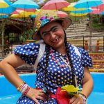 Una mujer con vestido azul de lunares y sombrero de paja posa alegremente junto a una piscina del Parque los Arrieros, rodeada de sombrillas de colores. Lleva el pelo trenzado, maquillaje brillante y sostiene una flor amarilla.