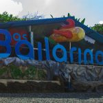 Grandes letras azules escriben "Bailarinos" con una escultura de un pez encima y vegetación de fondo, formando una colorida señal del Parque las Bailarinas.