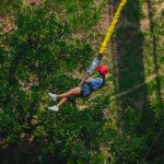 Una persona con casco y arnés rojos se lanza en tirolina por encima de los verdes árboles del Parque de las Bailarinas, con las piernas estiradas y los brazos sujetando el equipo, mientras la luz del sol proyecta sombras en el suelo.
