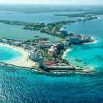 Vista aérea de una ciudad costera con playas de arena blanca, agua turquesa, hoteles de gran altura y una delgada franja de tierra que se curva hacia el océano. Los edificios están rodeados de mar por ambos lados bajo un cielo azul despejado.