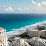 Vista aérea de hoteles y complejos turísticos en primera línea de playa que bordean una larga franja de playa de arena blanca con olas de color turquesa bajo un cielo parcialmente nublado.