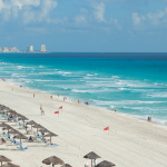 Amplia playa de arena y agua turquesa, hileras de sombrillas de paja, gente paseando por la orilla, hoteles y complejos turísticos bordeando la costa y un cielo parcialmente nublado.