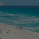 Una vista panorámica de una playa de arena con olas turquesas, hileras de sombrillas de paja, palmeras y lejanos rascacielos bajo un cielo azul despejado.
