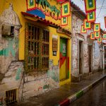 Una calle vibrante y colorida con edificios pintados, una ventana decorativa amarilla y verde, y banderas rojas, verdes y amarillas colgadas en lo alto. La calle está mojada, reflejo de la lluvia reciente.