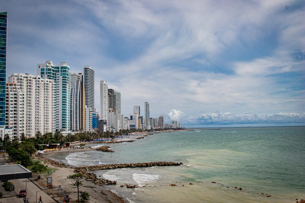 Un paisaje urbano costero con altos edificios modernos que bordean una playa de arena con palmeras, suaves olas en el mar turquesa y un cielo parcialmente nublado.