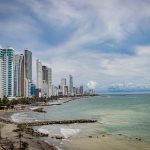 Un paisaje urbano costero con altos edificios modernos que bordean una playa de arena con palmeras, suaves olas en el mar turquesa y un cielo parcialmente nublado.
